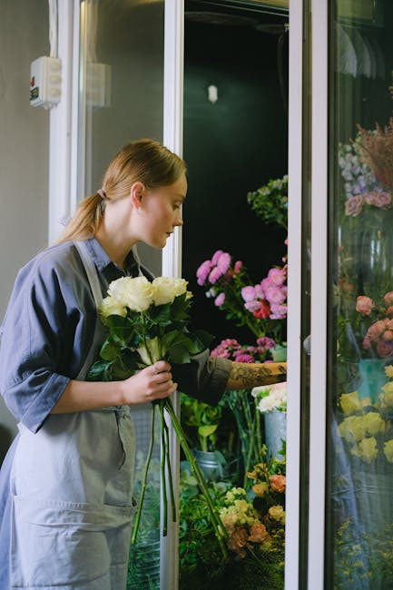 A young woman with light brown hair tied back, dressed in a blue shirt and light grey apron, selecting a bouquet of white roses from a display at a florist shop. The arrangement is tied with a dark gr