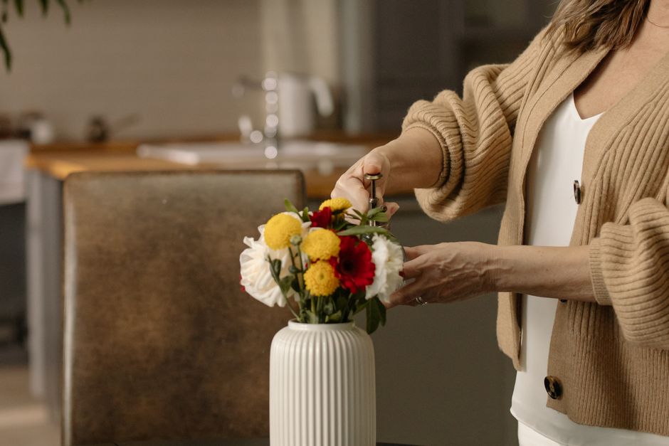 A person wearing a beige cardigan and white top arranging a floral bouquet in a tall, white, ribbed ceramic vase on a wooden table in a well-lit kitchen. The bouquet features a mix of fresh flowers in