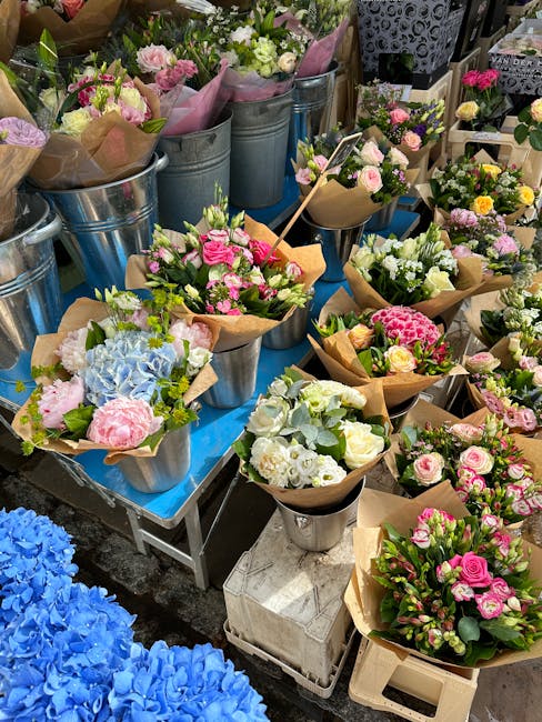 A display of freshly arranged flower bouquets featuring pink roses, white hydrangeas, and green foliage wrapped in brown kraft paper. The bouquets are placed in shiny metal buckets and small vases, ar