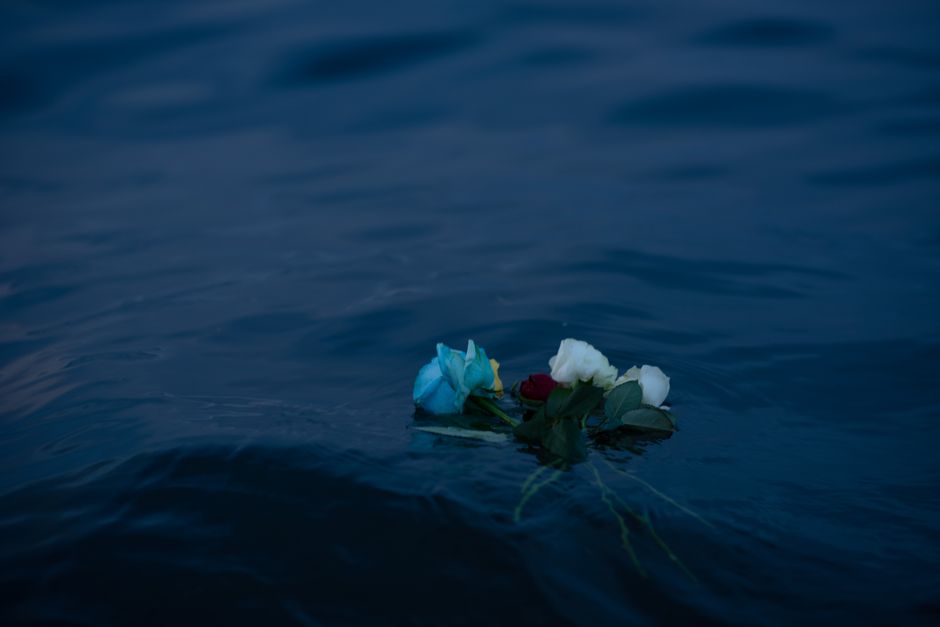 A close-up of a small bouquet of flowers floating on dark, rippling water. The arrangement includes a light blue rose, white roses, a single red rose, and green foliage, all with fresh, vibrant petals