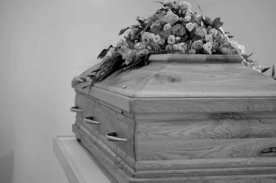A wooden casket with a smooth, polished surface and visible natural grain rests on a stand, topped with an elaborate floral tribute. The floral arrangement includes an assortment of fresh flowers such